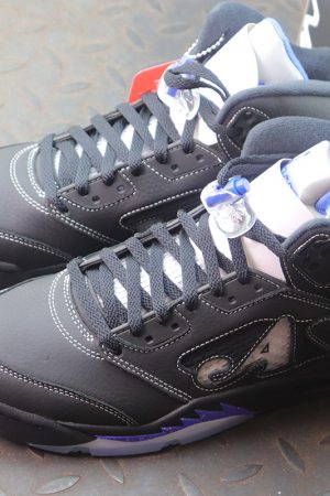 A pair of Awake NY x Air Jordan 5 sneakers in black with racer blue accents. The shoes feature a black leather upper, an "A" logo on the side mesh, and a blue speckled midsole. The sneakers are placed on a gray metal surface.