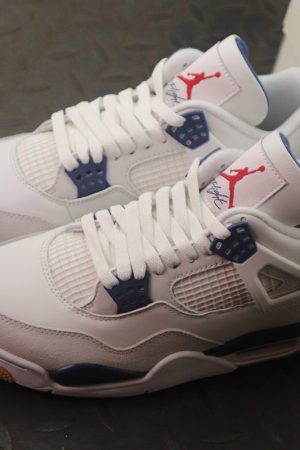 A pair of Nike SB x Air Jordan 4 sneakers. The shoes have a white leather upper, navy blue lace wings and heel tab, a red Jumpman logo on the tongue, and a gum-colored outsole.